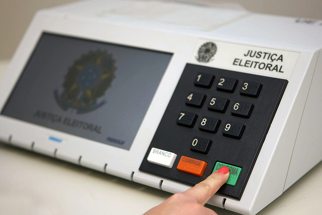 Urna eletrônica da Justiça Eleitoral, com um dedo pressionando a tecla verde confirma.